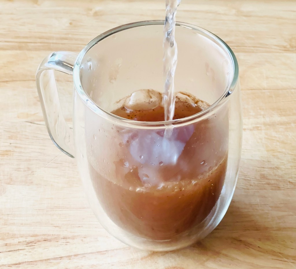 A clear mug with 1 cinnamon spice ice cube dissolving under boiling water