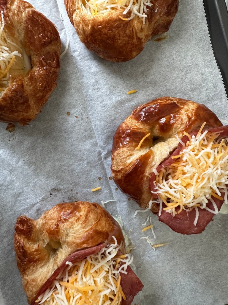 Croissants on a parchment paper-lined baking sheet, topped with slices of turkey bacon, eggs, and shredded Mexican cheese