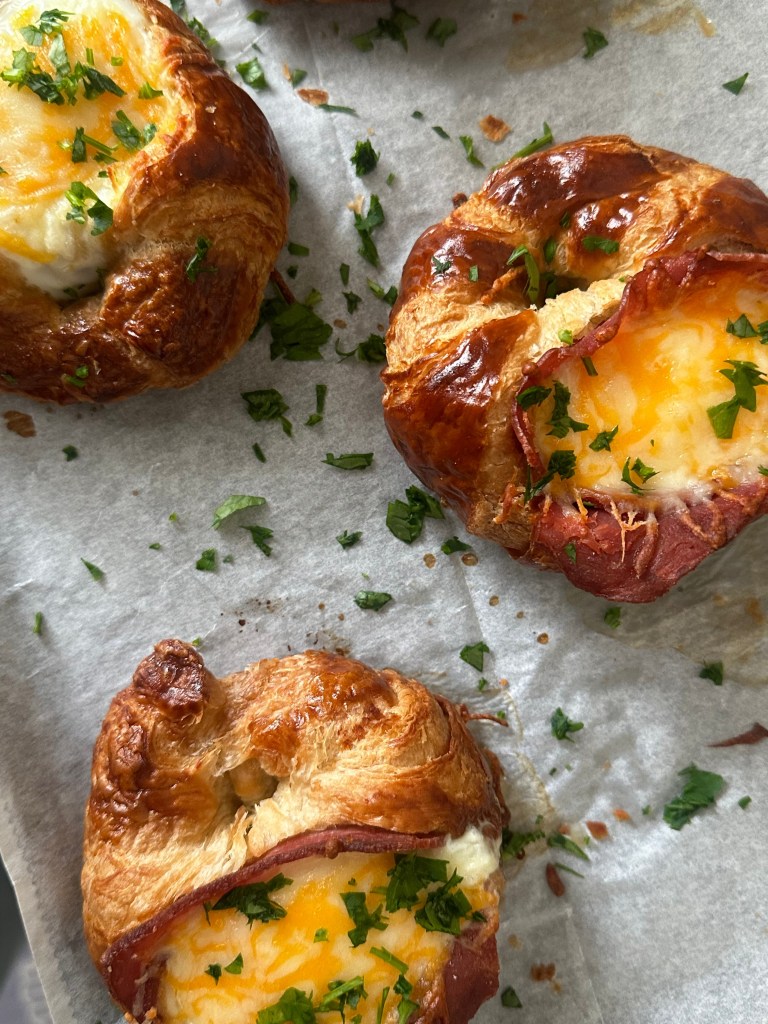 Baked croissants on a parchment paper-lined baking sheet, topped with slices of turkey bacon, eggs, and melted Mexican cheese and garnished with fresh parsley