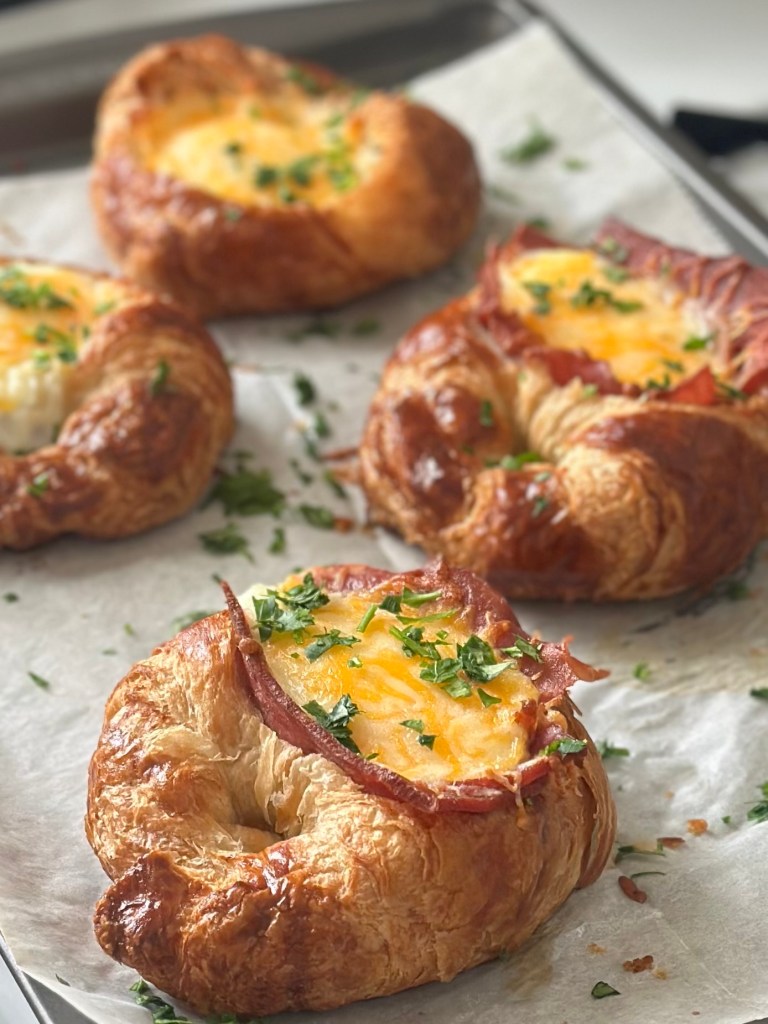 Baked croissants on a parchment paper-lined baking sheet, topped with slices of turkey bacon, eggs, and melted Mexican cheese and garnished with fresh parsley