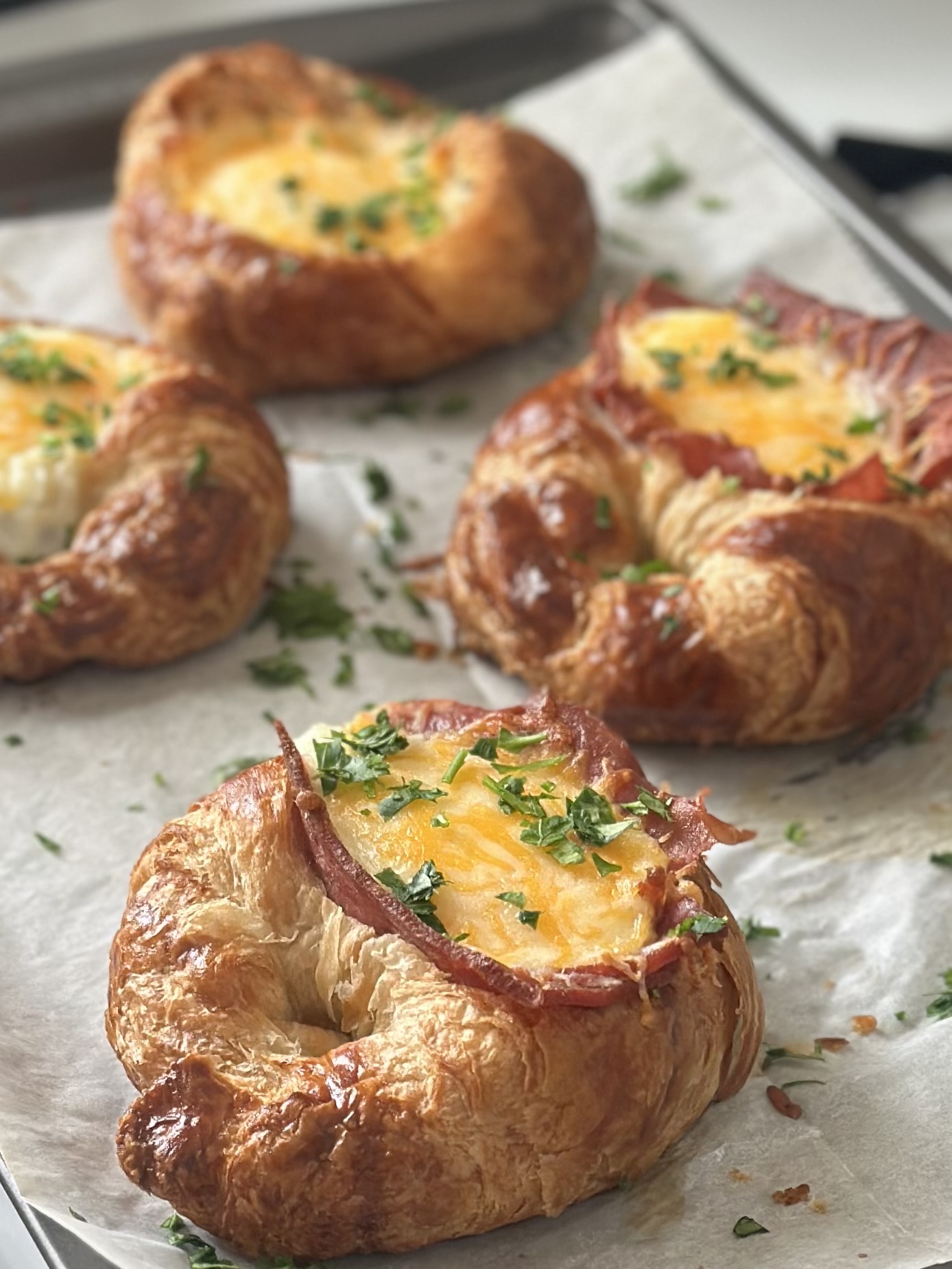 Baked croissants on a parchment paper-lined baking sheet, topped with slices of turkey bacon, eggs, and melted Mexican cheese and garnished with fresh parsley