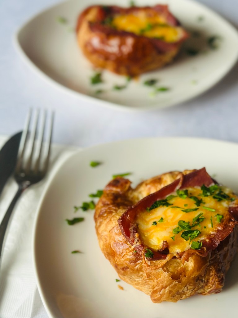 A serving plate of baked croissants, topped with slices of turkey bacon, eggs, and melted Mexican cheese and garnished with fresh parsley