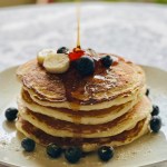 Stack of pancakes on a plate with bananas, blueberries, and syrup being drizzled on top