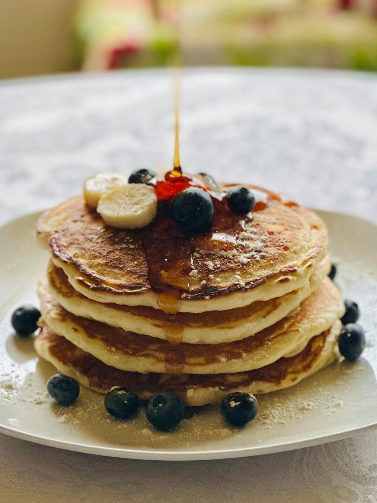Stack of pancakes on a plate with bananas, blueberries, and syrup being drizzled on top