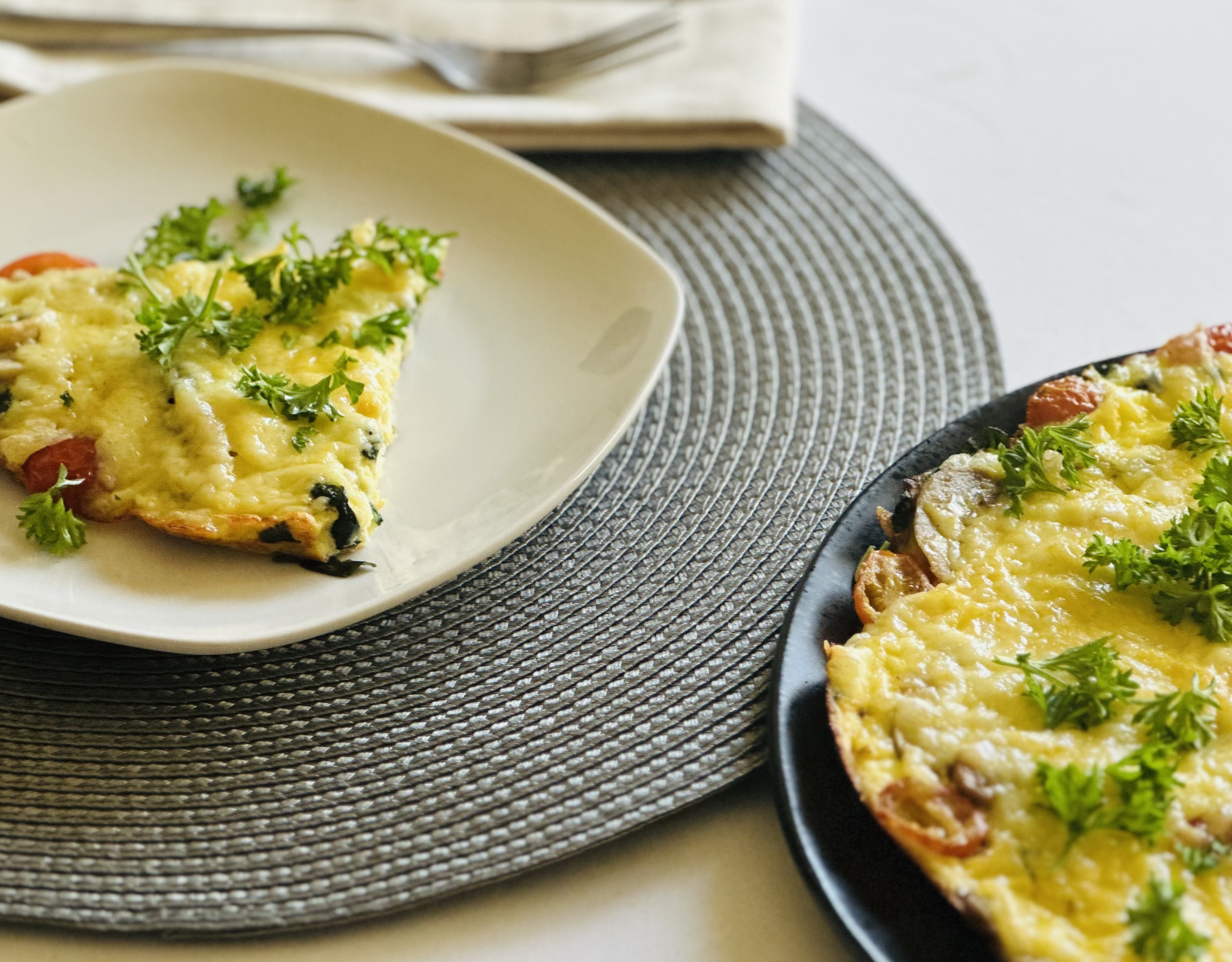 A slice of homemade egg frittata topped with fresh parsley, served on a white plate alongside a whole frittata on a black plate.