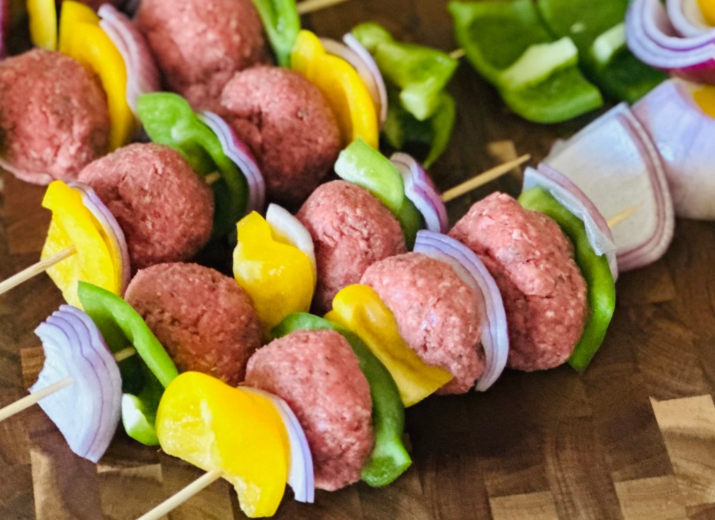 A close-up image of meatball kebabs assembled on wooden skewers, featuring alternating pieces of raw beef meatballs, yellow bell peppers, green bell peppers, and red onions, placed on a wooden cutting board.