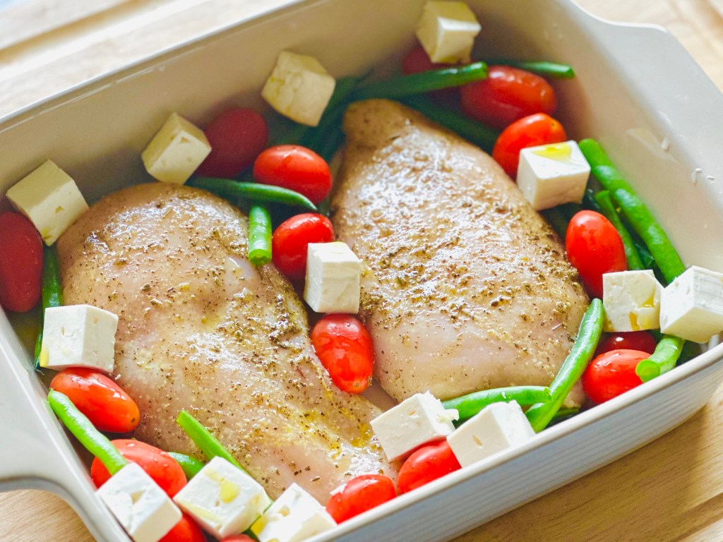 Baking dish with uncooked chicken, tomatoes, green beans, and feta