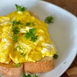 A slice of toasted bread with scrambled eggs and parsley on top being served at a table