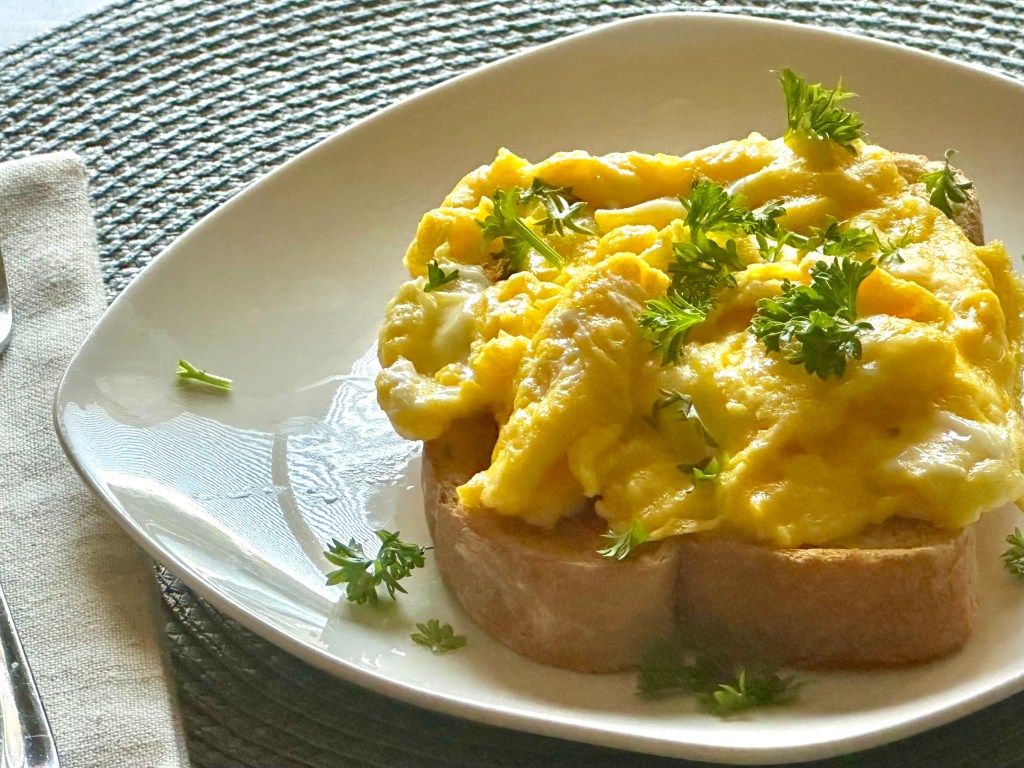 A slice of toasted bread with scrambled eggs and parsley on top being served at a table