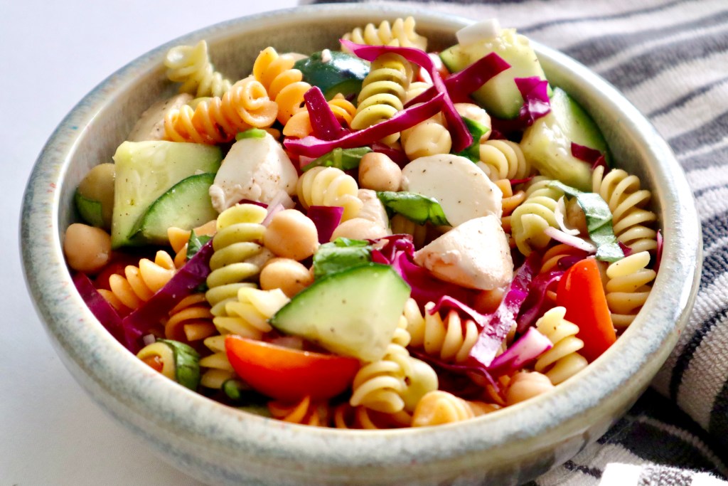 A vibrant bowl of easy pasta salad featuring rotini pasta, colorful vegetables, chickpeas, and mozzarella, served in a ceramic bowl with a striped cloth in the background.