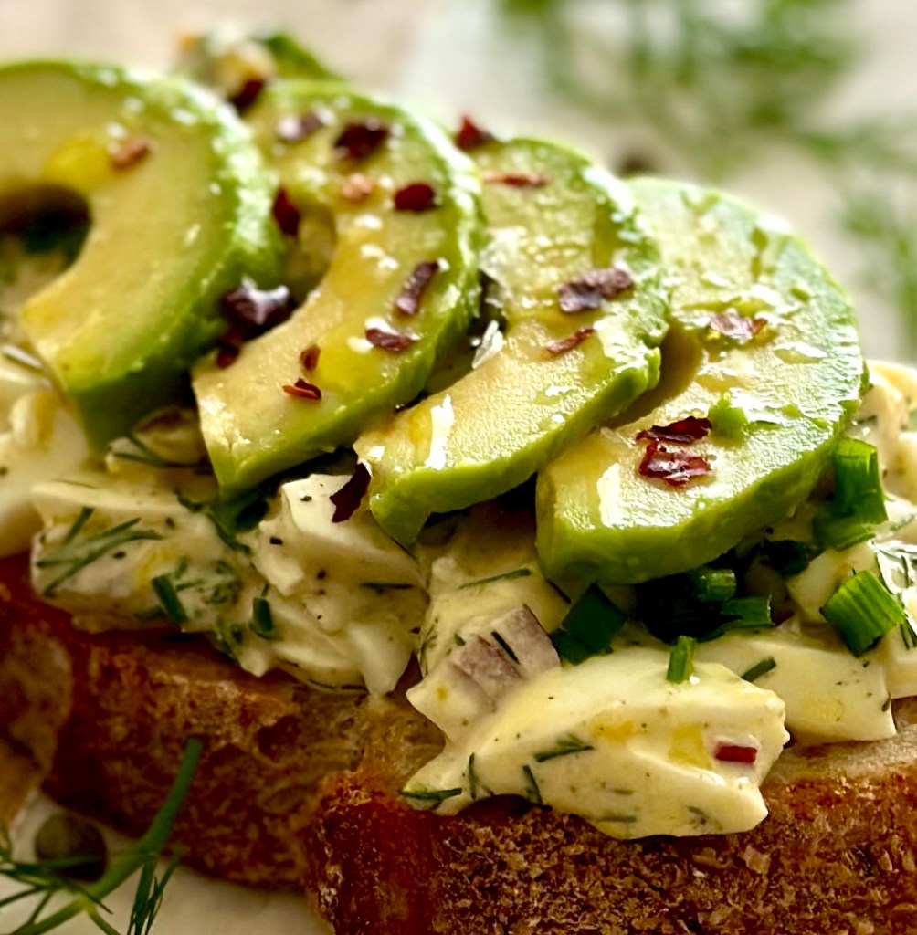 A slice of sourdough bread with egg salad and avocado slices on top