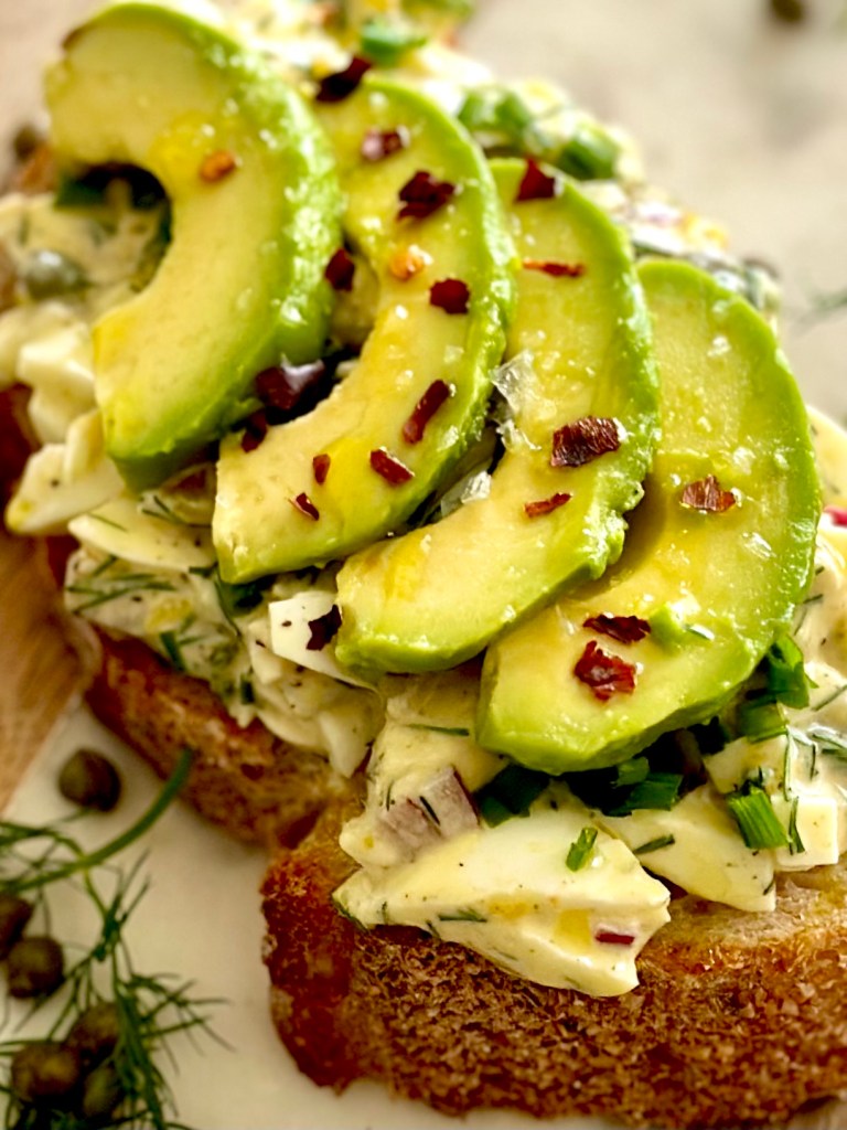 A slice of sourdough bread with egg salad and avocado slices on top