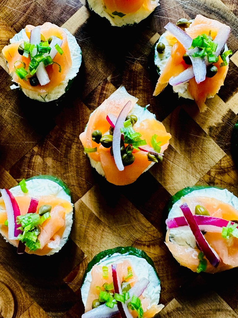 A wooden board full of smoked salmon, red onion, and capers on a bed of cream cheese atop cucumber slices