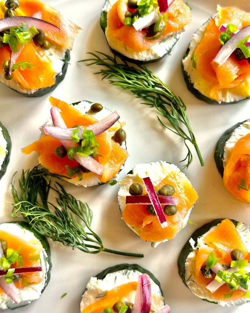 A serving plate full of smoked salmon, red onion, and capers on a bed of cream cheese atop cucumber slices