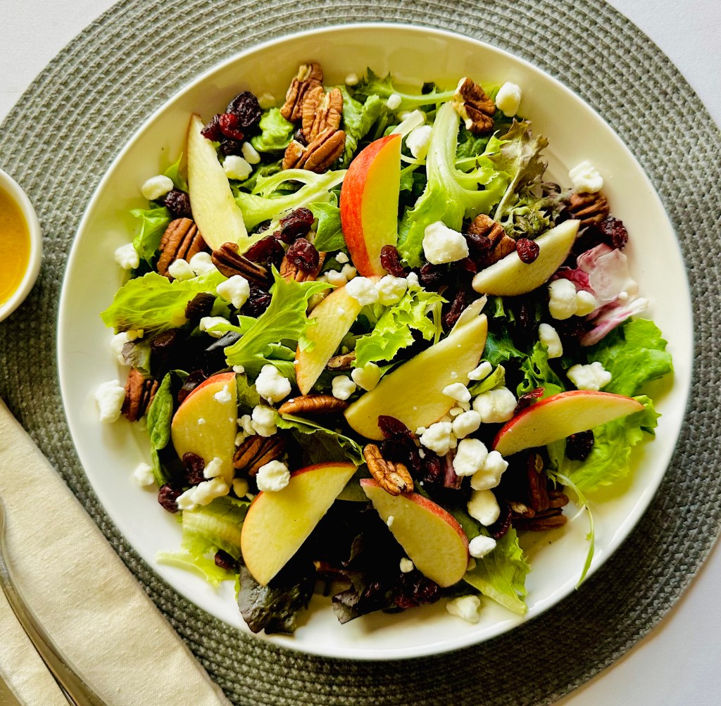 An autumn salad with apple slices, dried cranberries, crumbled goat cheese, and pecans, served on a serving plate