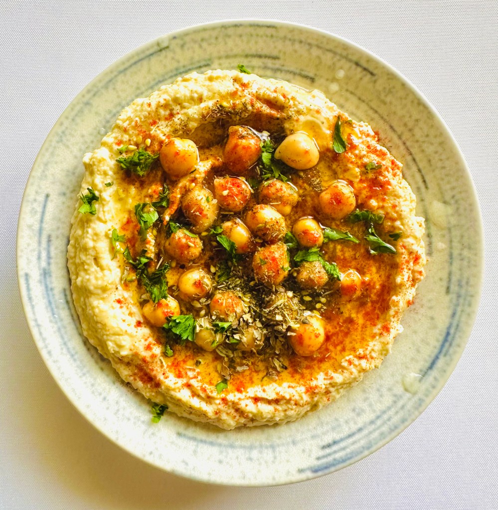 A bowl of homemade hummus topped with chickpeas, olive oil, parsley, and spices, served on a patterned plate.