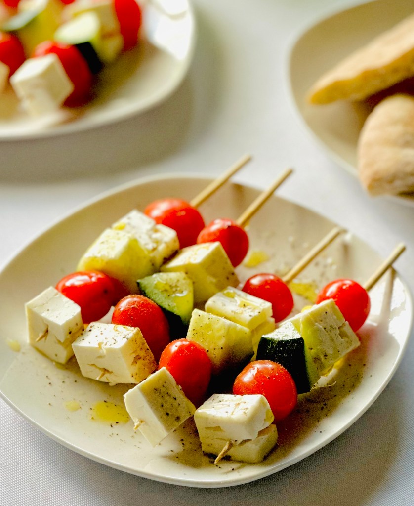 A plate with four skewers threaded with cherry tomatoes, cubes of feta, and cucumber