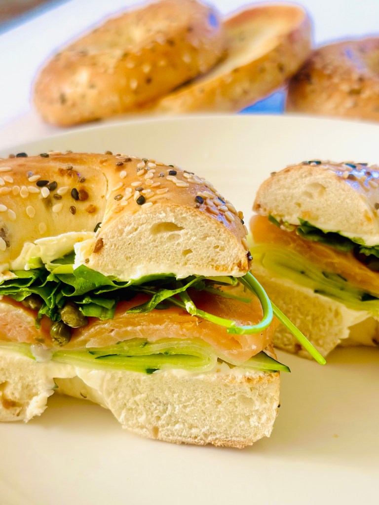 A sliced bagel sandwich with cream cheese, cucumber, smoked salmon, capers, and arugula
