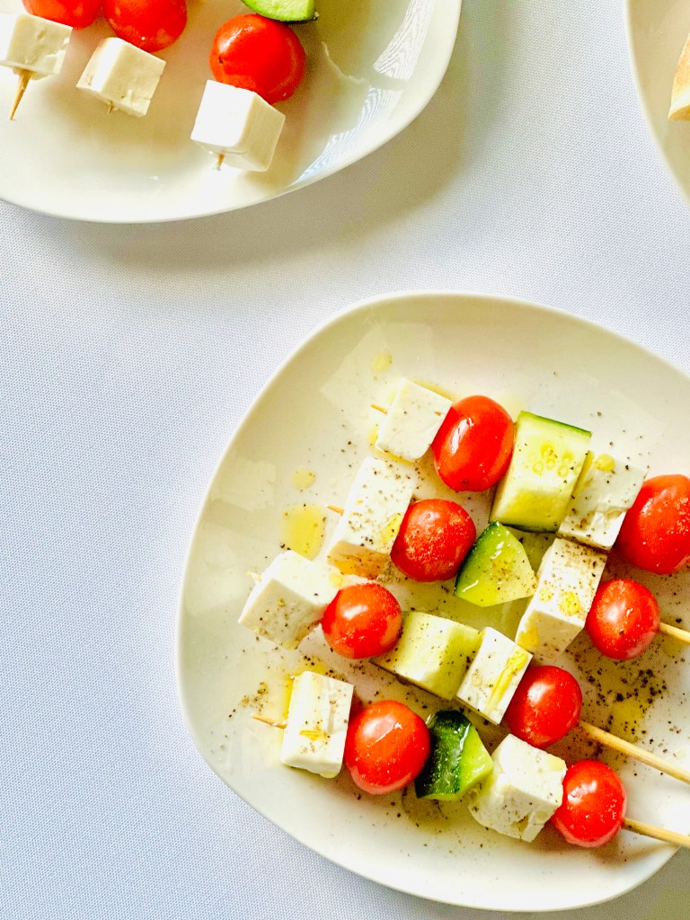 A plate with four skewers threaded with cherry tomatoes, cubes of feta, and cucumber