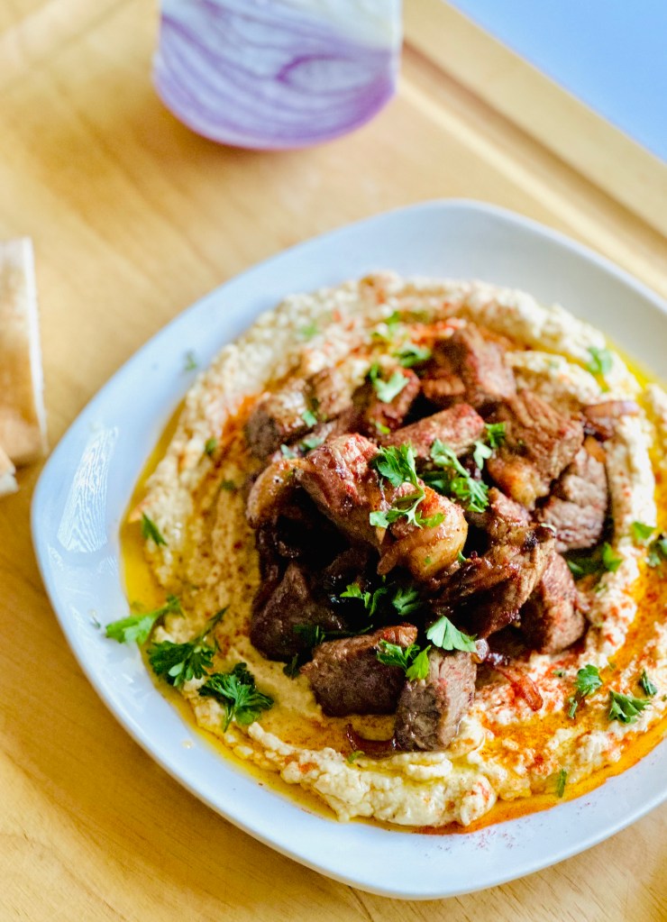 Homemade hummus topped with sirloin steak on a plate, garnished with freshly chopped parsley
