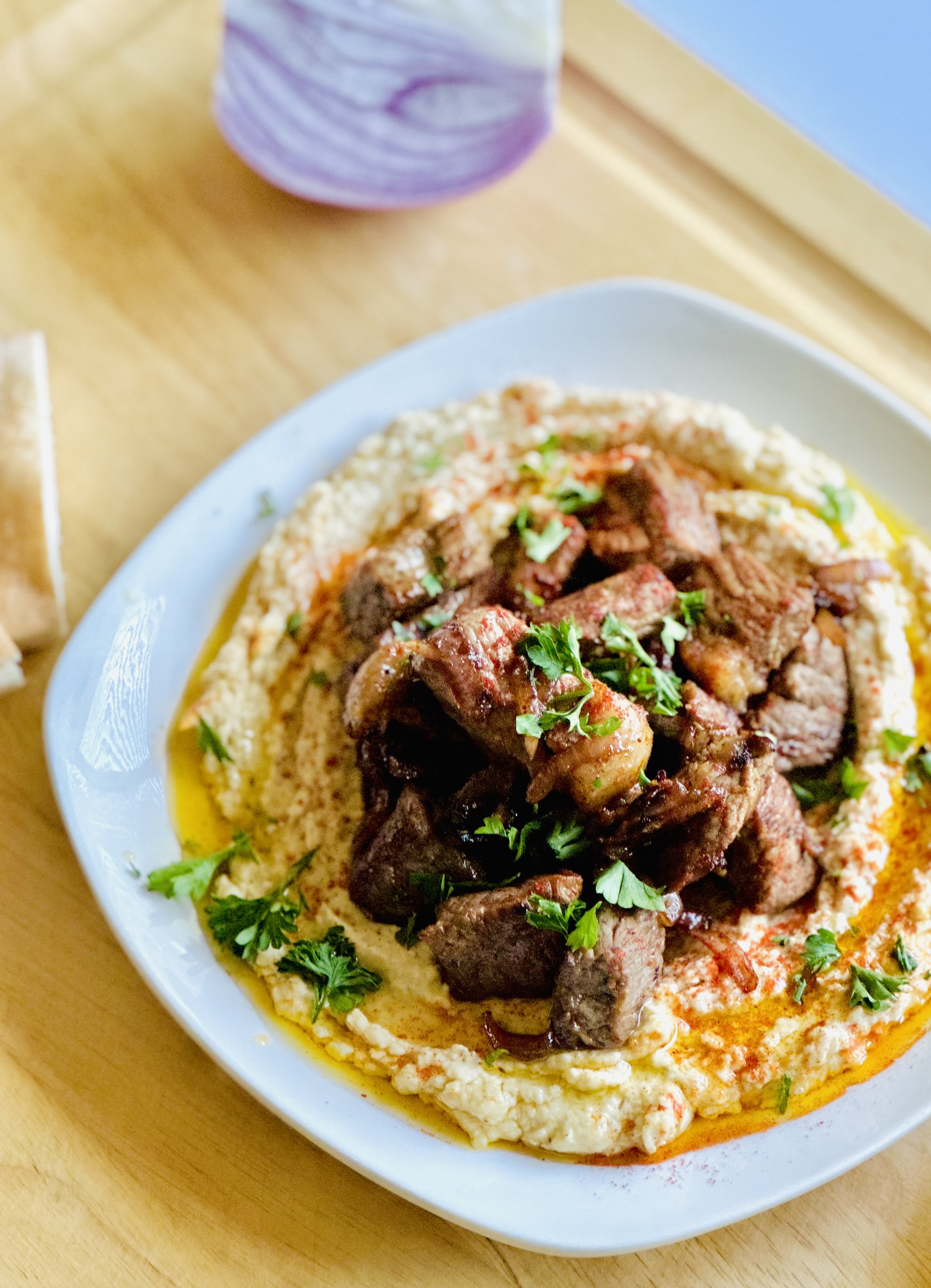 Homemade hummus topped with sirloin steak on a plate, garnished with freshly chopped parsley