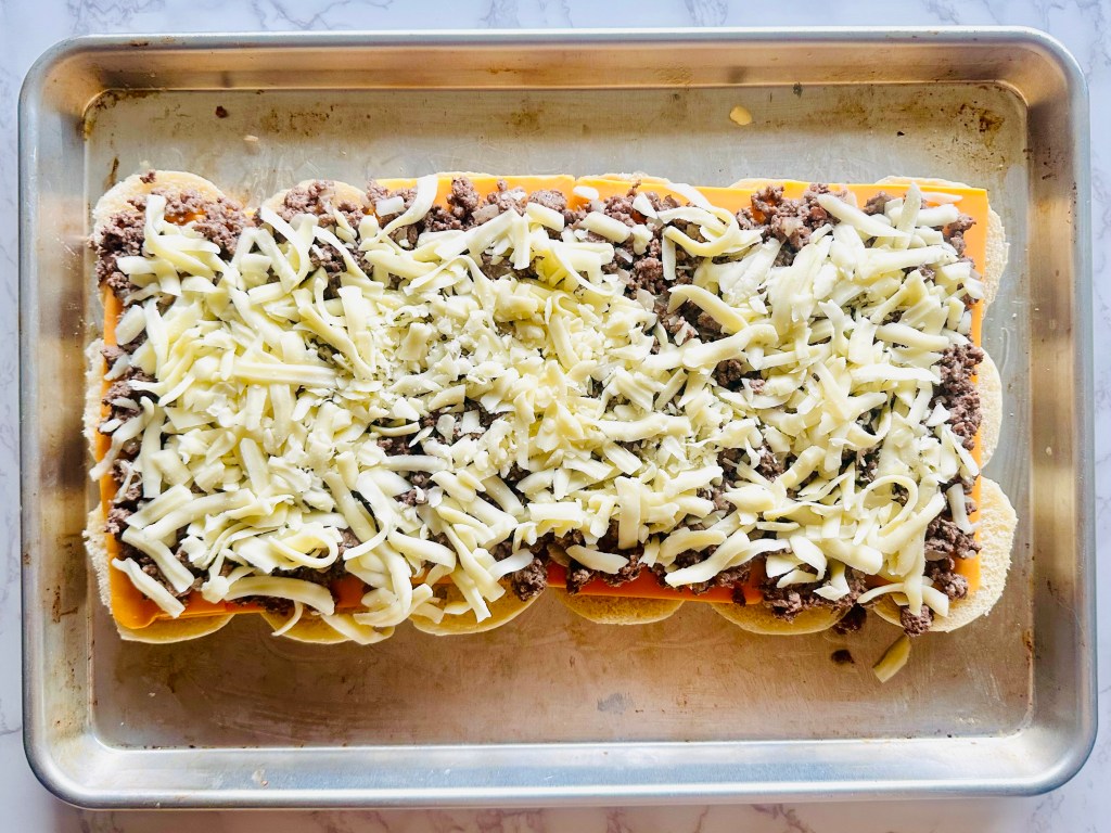 Slider buns being prepared with sliced cheddar cheese, ground beef, and shredded cheddar