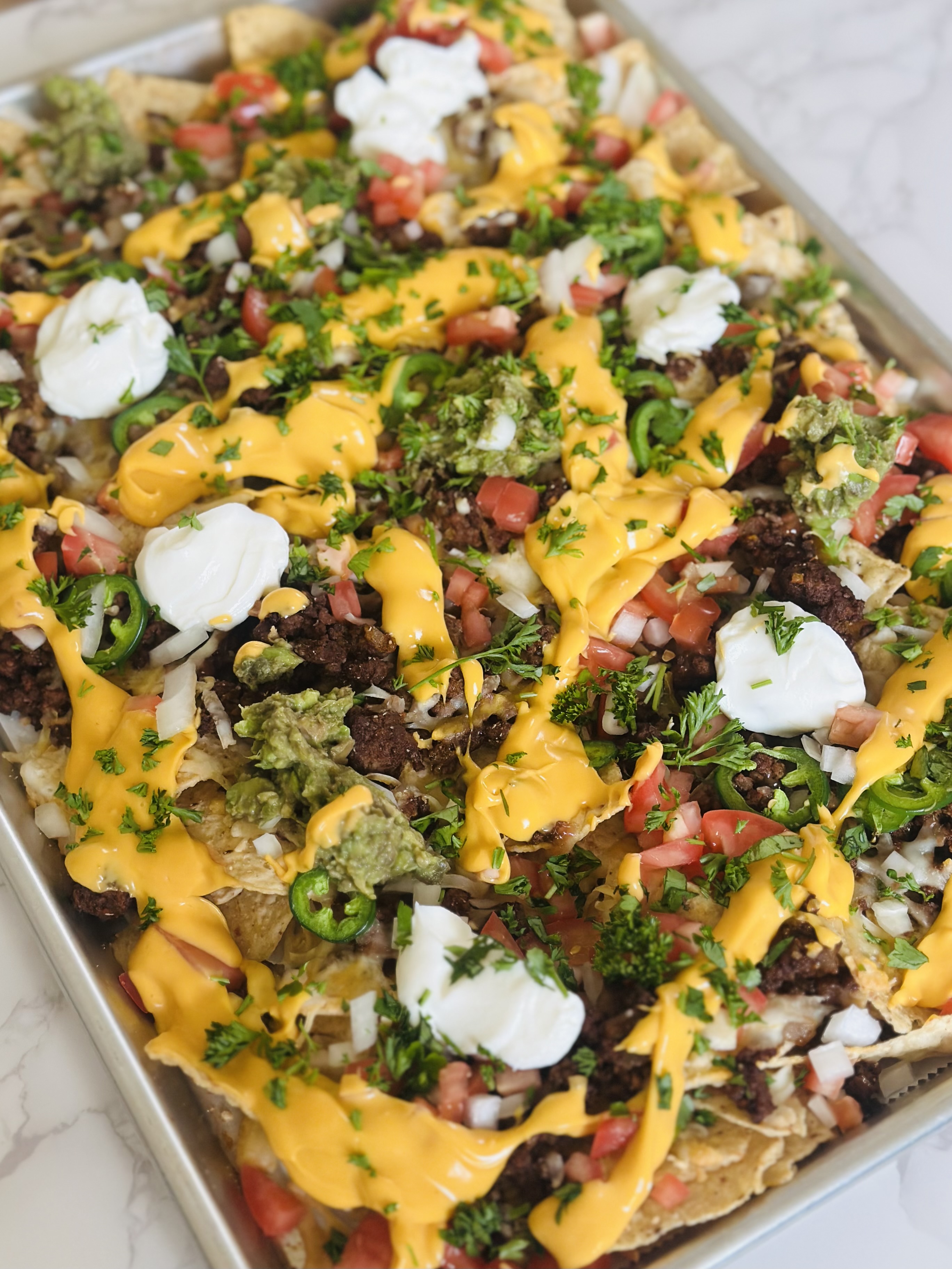 A sheet pan laid out with tortilla chips, ground beef, melted Mexican-blend cheese and nacho cheese sauce, and jalapenos
