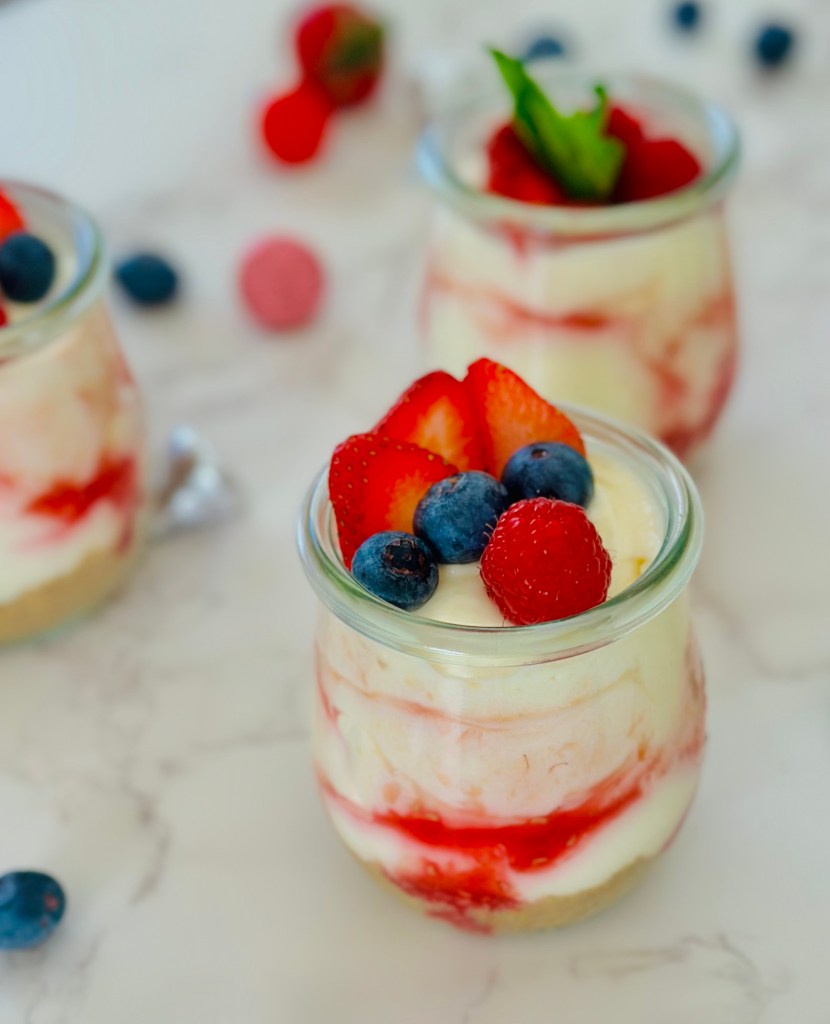 A photo of no-bake cheesecake cups with raspberry sauce, fresh berry toppings, and mint leaves