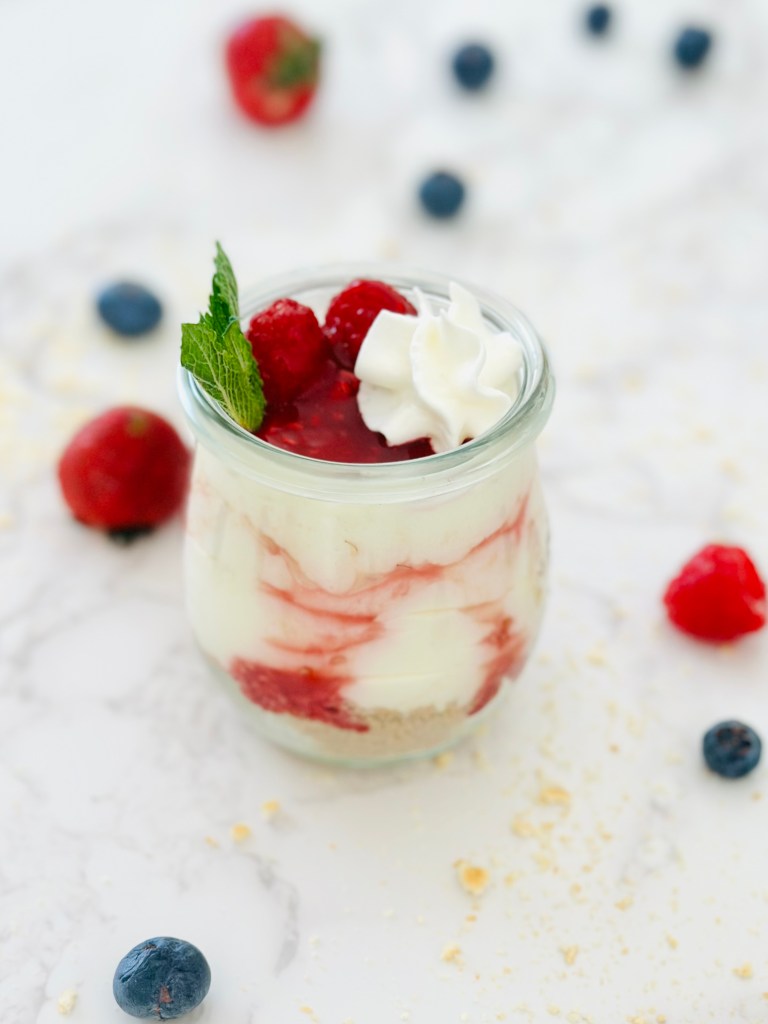 A no-bake cheesecake cup with graham cracker crust, cream filling, and a topping of raspberries, mint leaves, and whipped cream
