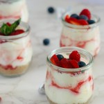 A photo of no-bake cheesecake cups with raspberry sauce, fresh berry toppings, and mint leaves