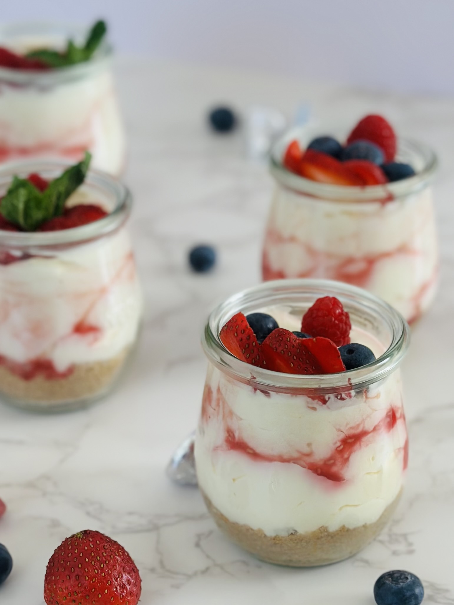 A photo of no-bake cheesecake cups with raspberry sauce, fresh berry toppings, and mint leaves