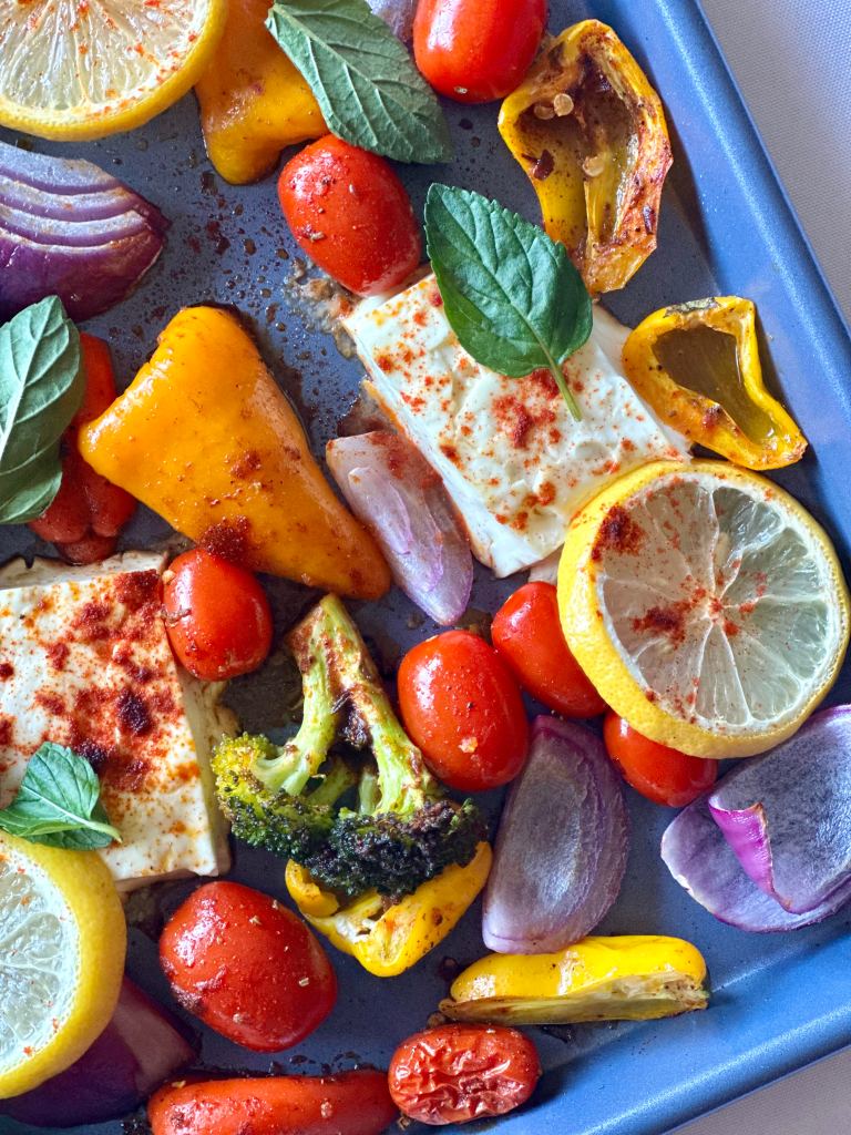 A sheet pan filled with broccoli, tomatoes, red onion, and sweet peppers that are seasoned and layered with feta cheese and lemon slices