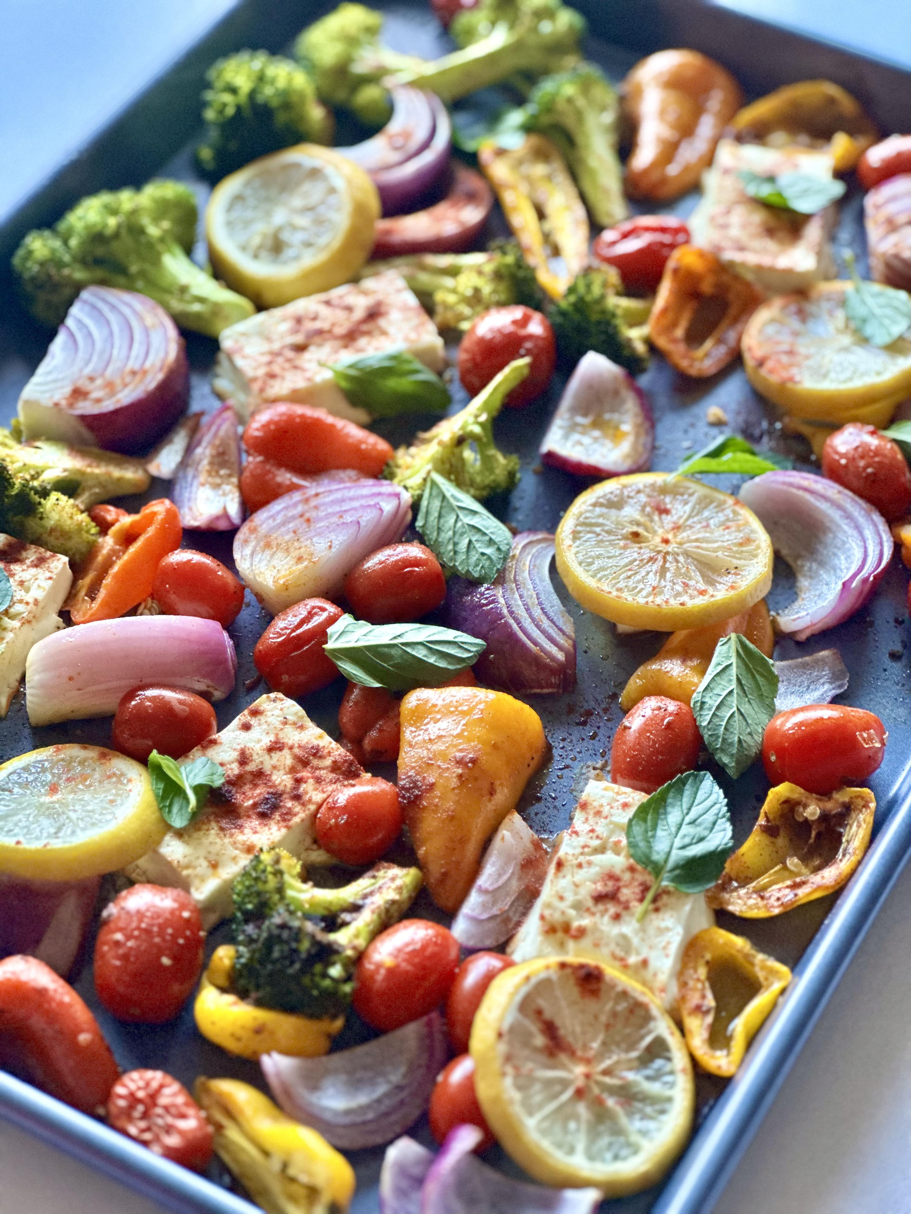 A sheet pan filled with broccoli, tomatoes, red onion, and sweet peppers that are seasoned and layered with feta cheese and lemon slices
