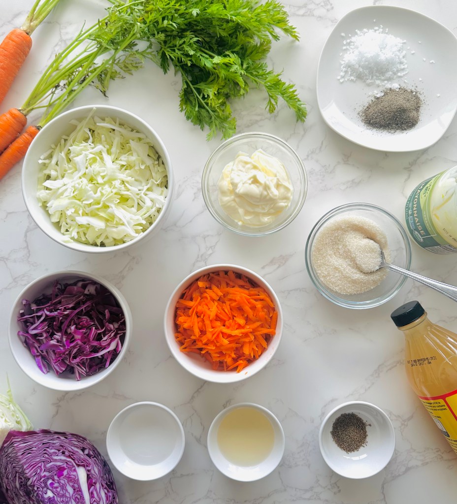 A photo of ingredients used to make homemade coleslaw with dressing: shredded cabbage, shredded carrots, mayo, white and apple cider vinegars, sugar, celery seed, salt, and pepper
