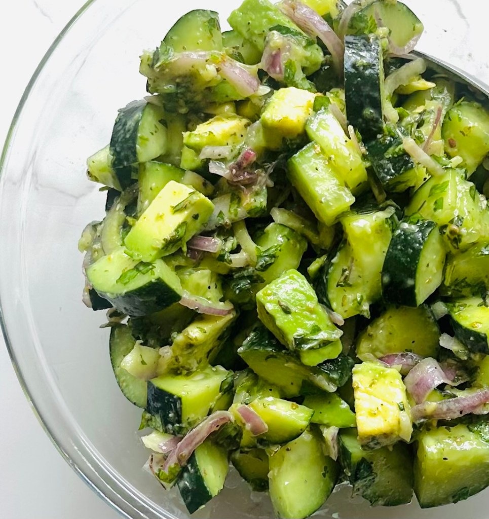 A glass bowl with cucumber, avocado, red onion, chopped basil and cilantro, Italian seasoning, and olive oil being tossed into a salad