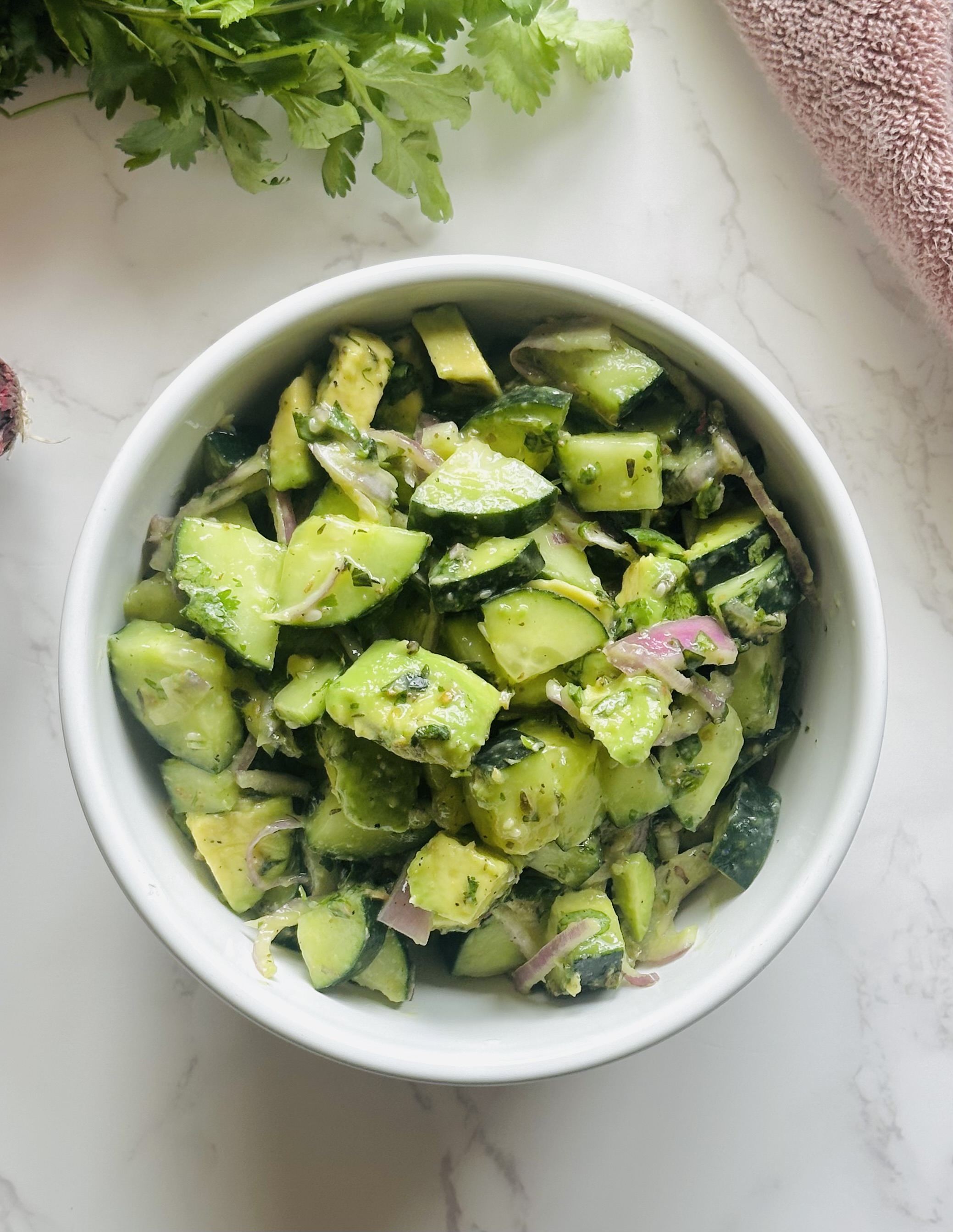 A medium salad bowl full of cucumber, avocado, red onion, chopped basil and cilantro, Italian seasoning, olive oil, salt, and pepper