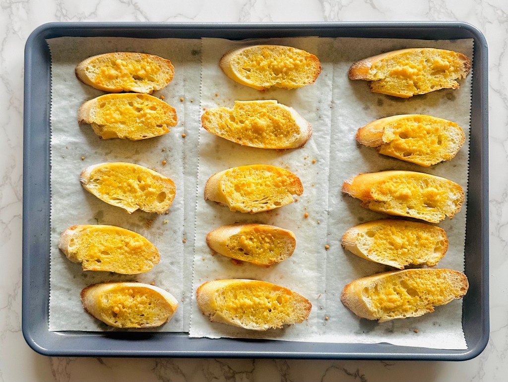 Baguette slices on a baking sheet that have been baked in the oven and brushed with olive oil and a sprinkle of grated Parmesan