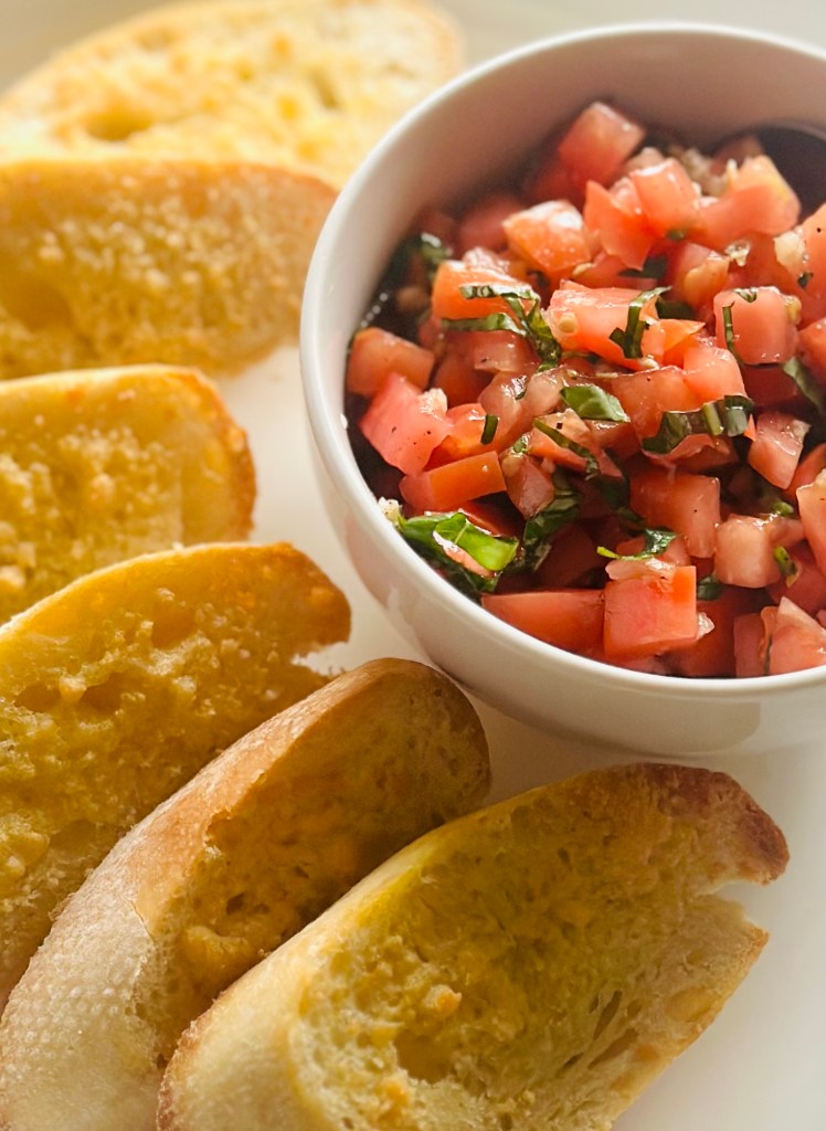 A platter with toasted baguette slices and a bowl of bruschetta topping made with Roma tomatoes, basil, garlic, balsamic vinegar, olive oil, salt, and pepper