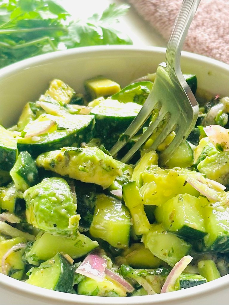 A fork reaching into a salad bowl of cucumber, avocado, red onion, chopped basil and cilantro, Italian seasoning, olive oil, salt, and pepper