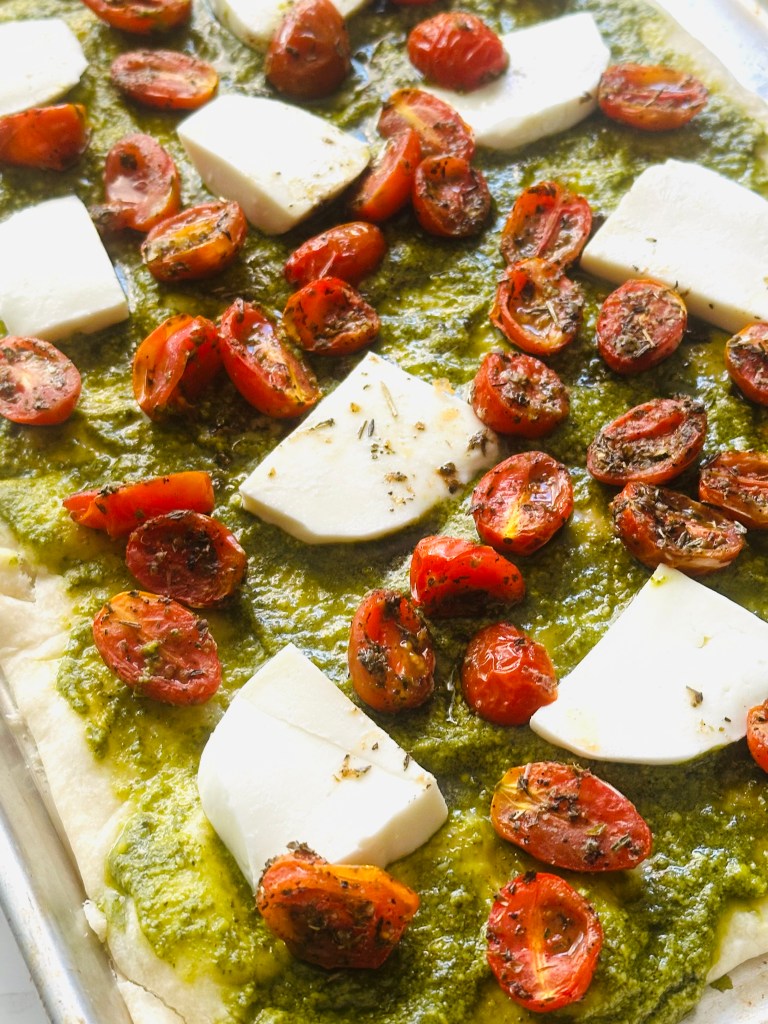 A sheet pan lined with Jus-Rol pizza crust dough, topped with pesto, buffalo mozzarella, and roasted tomatoes