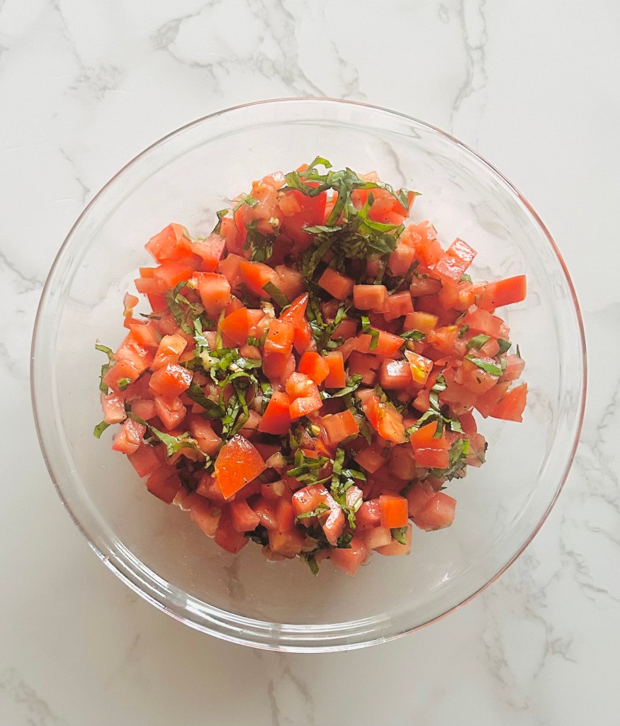 A glass bowl containing bruschetta topping made with diced Roma tomatoes, basil, garlic, balsamic vinegar, olive oil, salt, and pepper