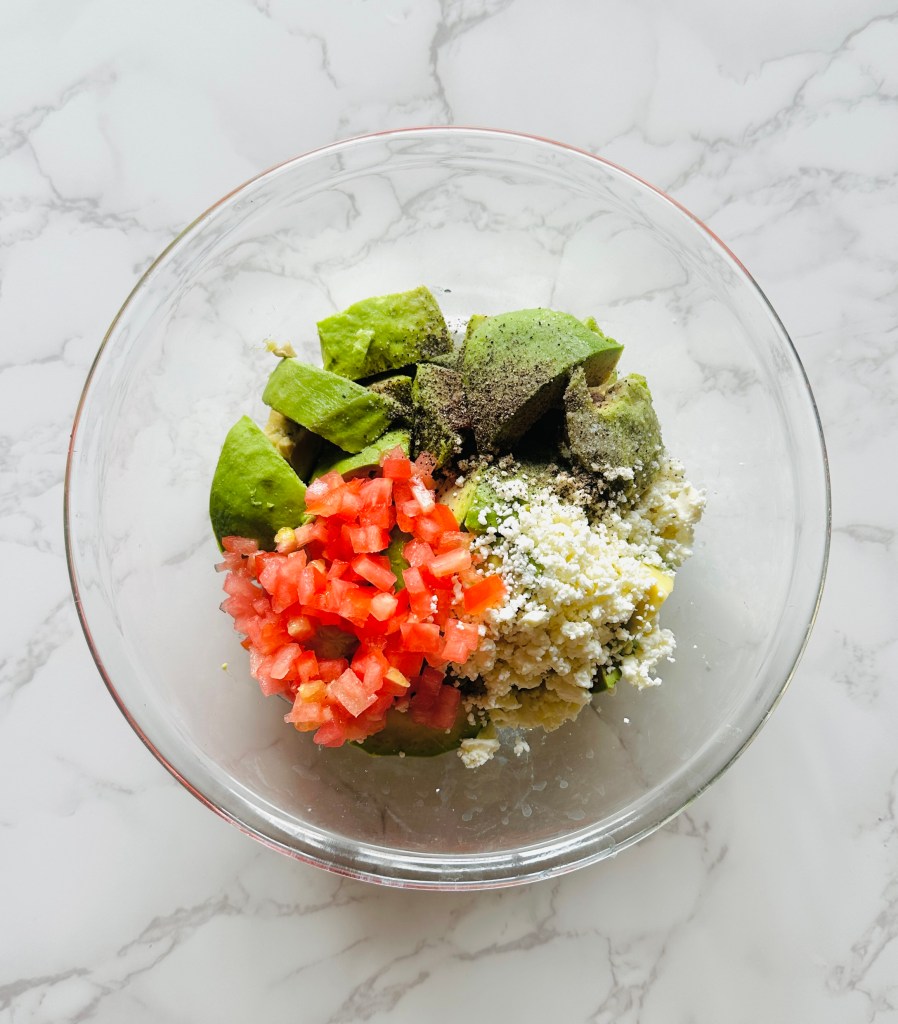 A glass bowl containing ingredients for avocado mash: avocado, crumbled feta, lemon juice, tomato, salt, and pepper