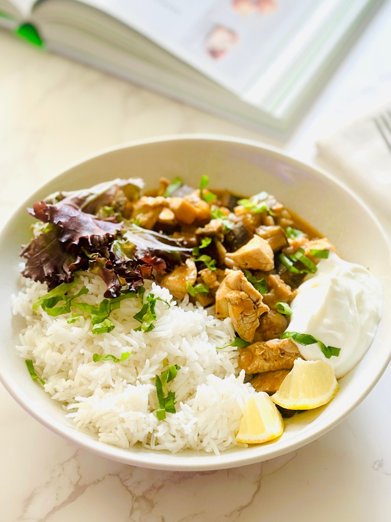 A serving plate of chicken eggplant curry served with a dollop of Greek yogurt on the side, lemon wedges, handful of mixed greens, and jasmine rice