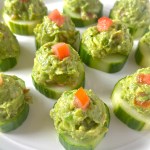 A serving platter filled with sliced cucumber rounds that are topped with an avocado mash of avocados, tomatoes, feta, lemon juice, salt, and pepper