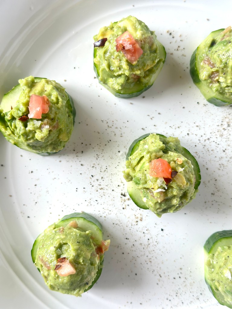 A serving platter filled with sliced cucumber rounds that are topped with an avocado mash of avocados, tomatoes, feta, lemon juice, salt, and pepper