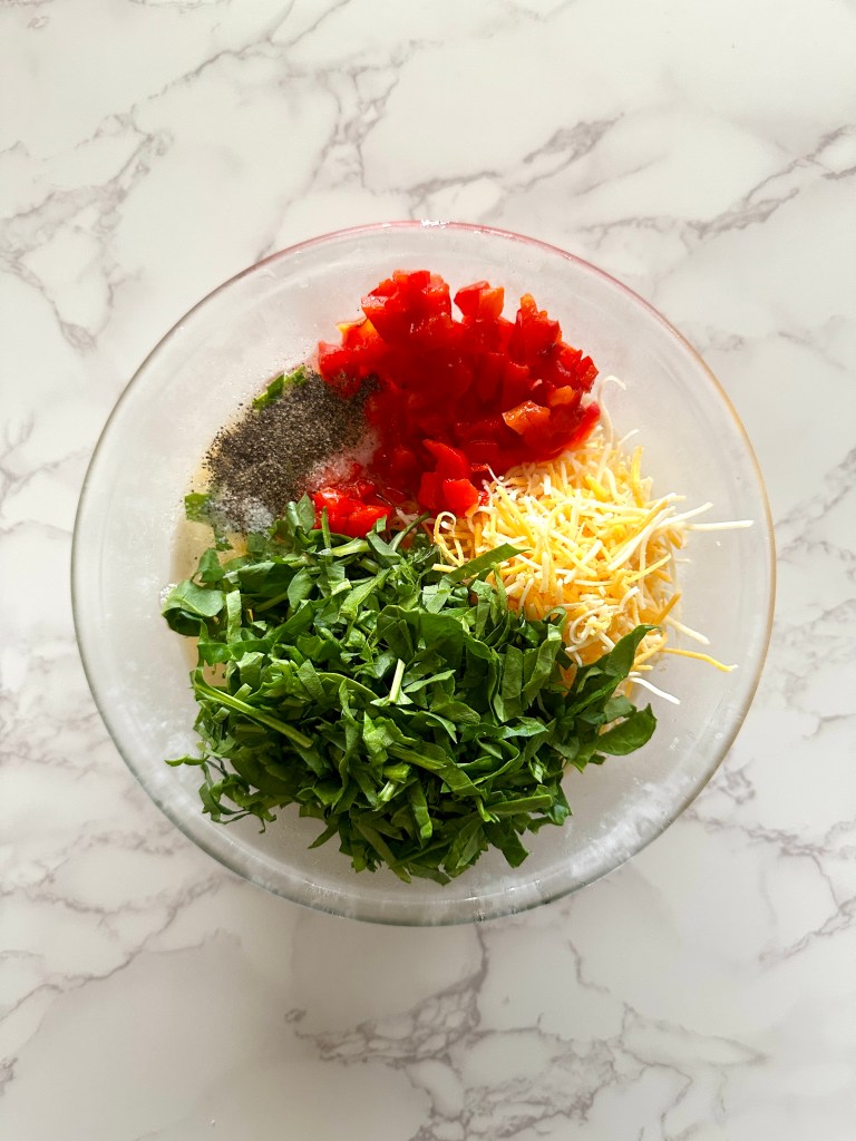 A clear glass bowl full of egg yolks, chopped spinach, shredded Mexican cheese, roasted red pepper, and salt and pepper