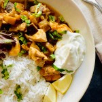 A serving plate of chicken eggplant curry served with a dollop of Greek yogurt on the side, lemon wedges, handful of mixed greens, and jasmine rice