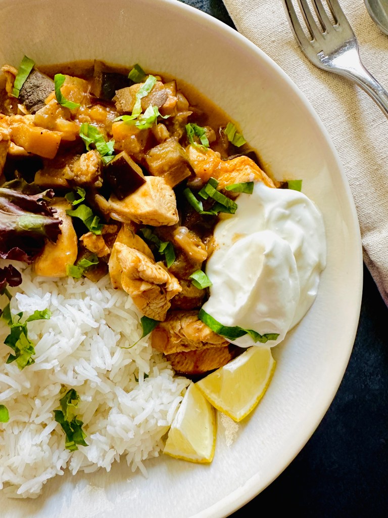 A serving plate of chicken eggplant curry served with a dollop of Greek yogurt on the side, lemon wedges, handful of mixed greens, and jasmine rice