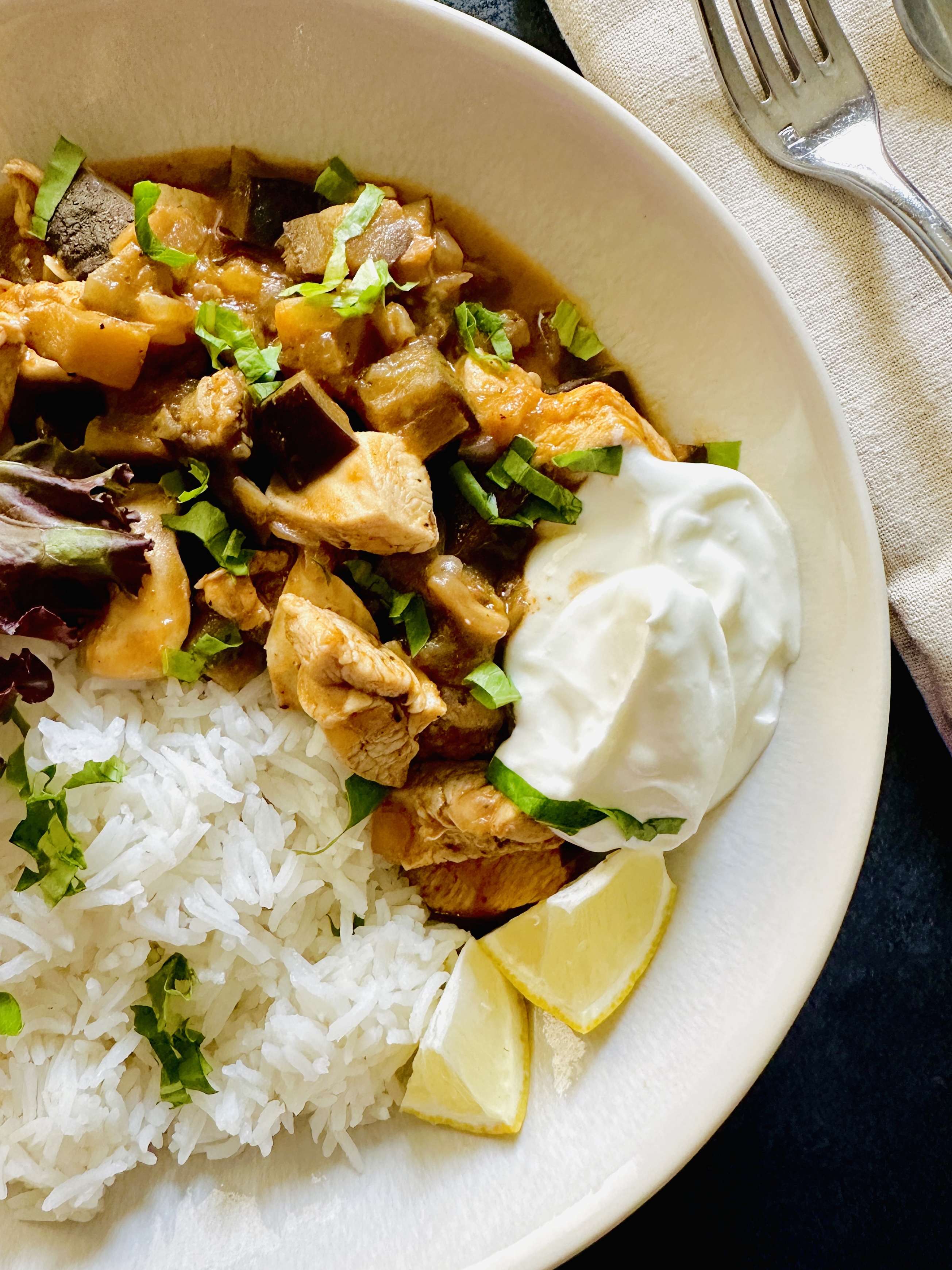 A serving plate of chicken eggplant curry served with a dollop of Greek yogurt on the side, lemon wedges, handful of mixed greens, and jasmine rice
