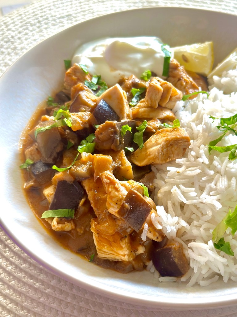A serving plate of chicken eggplant curry served with a dollop of Greek yogurt on the side, lemon wedges, and jasmine rice